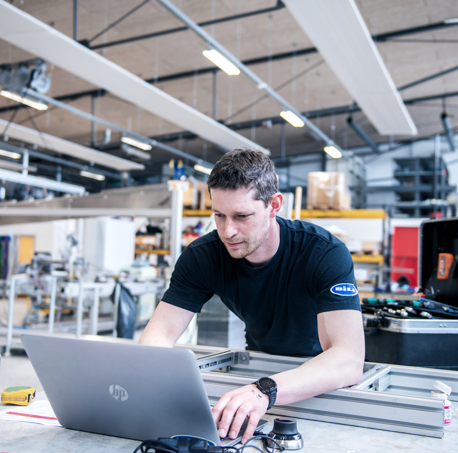 Mand i sort BILA-T-shirt arbejder på sin bærbare computer i et industrielt automatiseringsmiljø.